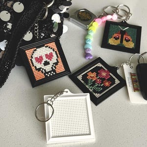 May include: Four square keychains with metal rings. One features a skull with heart eyes, one a butterfly, one a floral pattern, and one is a blank white grid. A small metal measuring tape and a pastel heart-shaped bead chain are included.