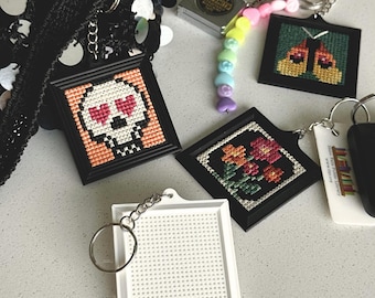 May include: Four square keychains with metal rings. One features a skull with heart eyes, one a butterfly, one a floral pattern, and one is a blank white grid. A small metal measuring tape and a pastel heart-shaped bead chain are included.