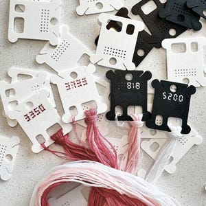 May include: A collection of white and black plastic embroidery floss bobbins featuring various numerical codes printed on the surface. Several bobbins are wound with pink, red, and white embroidery thread, while others remain empty.