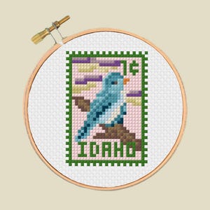 Puede incluir: Bordado de punto de cruz de un pájaro azul en una rama, que se asemeja a un sello postal. El diseño incluye la palabra "IDAHO" y una marca de "1¢". La obra de arte está enmarcada en un bastidor de bordado de madera.