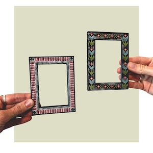 May include: Two rectangular frames featuring cross-stitch embroidery. The left frame has a red and white geometric pattern on a black base. The right frame displays a black base with a repeating floral motif in blue, green, and red thread.