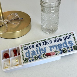 May include: A white rectangular pill organizer with a sliding lid featuring a cross-stitch pattern that reads Give us this day our daily meds in blue and green thread. The open compartments contain various red, white, and yellow pills.