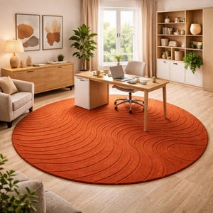 May include: A round, burnt orange rug with a wave pattern dominates a home office. The rug is centered in the room, with a wooden desk, chair, and a laptop. A beige armchair and wooden cabinet are also visible.