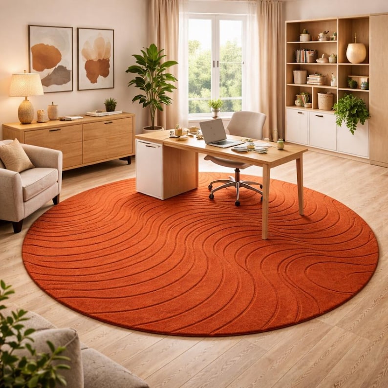 May include: A round, burnt orange rug with a wave pattern dominates a home office. The rug is centered in the room, with a wooden desk, chair, and a laptop. A beige armchair and wooden cabinet are also visible.