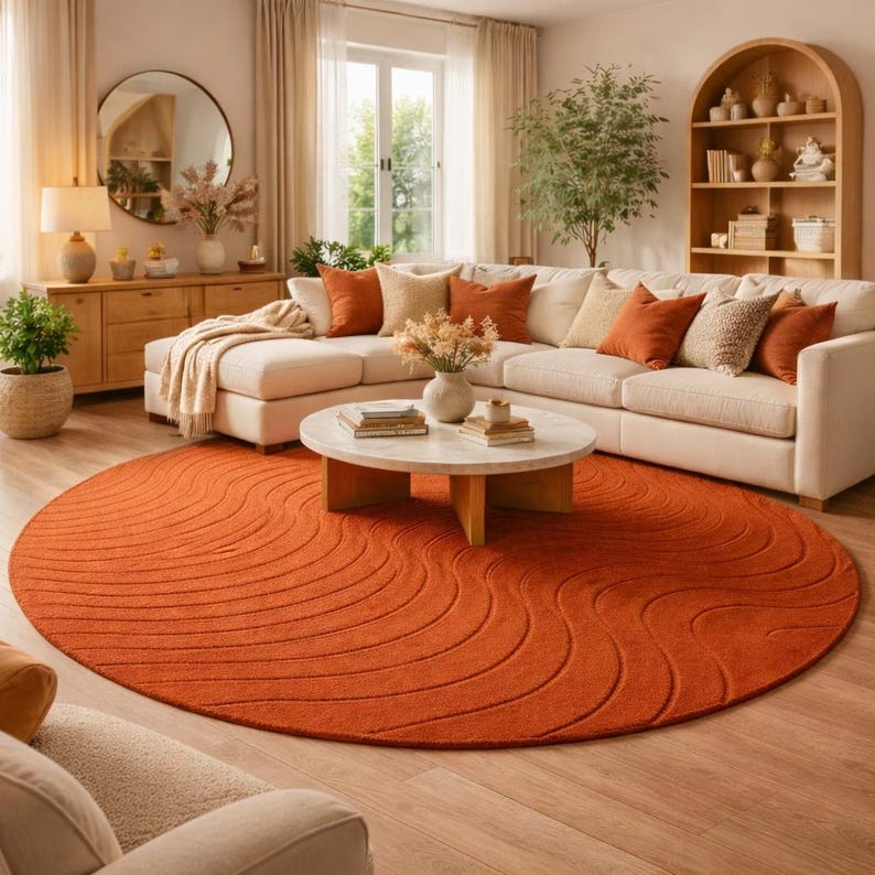 May include: A living room with a large, round, burnt orange rug with a wave pattern. A white sectional sofa with orange and beige pillows sits on the rug. A round marble coffee table and wooden furniture complete the room.