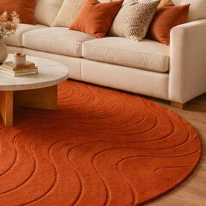 May include: A round, burnt orange rug with a wave pattern is in front of a cream-colored sofa with matching throw pillows. A light-colored coffee table with a marble top and wooden legs is in the foreground.