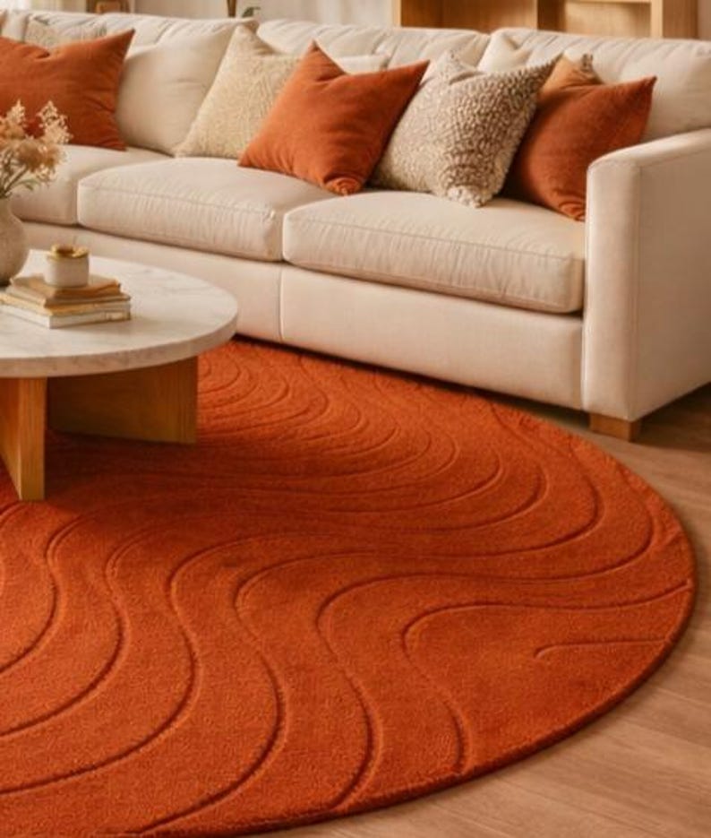 May include: A round, burnt orange rug with a wave pattern is in front of a cream-colored sofa with matching throw pillows. A light-colored coffee table with a marble top and wooden legs is in the foreground.