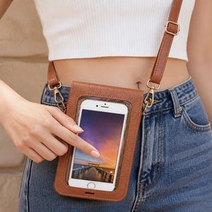 May include: A brown faux leather crossbody phone pouch with a clear plastic front window, featuring a smartphone inside. The pouch includes a thin, adjustable brown strap with gold-toned metal hardware, designed to be worn over the shoulder or across the body.