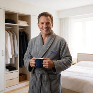 May include: A gray fleece bathrobe featuring a shawl collar, a tie waist, and the word Dad embroidered in blue thread on the left chest. The robe is worn with long sleeves and includes two front pockets, paired with a solid dark blue ceramic mug.