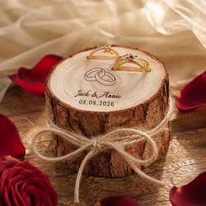 Peut inclure: Un porte-alliances cylindrique en bois avec des bords en écorce naturelle, noué avec un ruban en jute. La surface supérieure présente une gravure de deux anneaux entrelacés, les noms Jack & Annie et la date 08.08.2026, portant deux bagues en or.