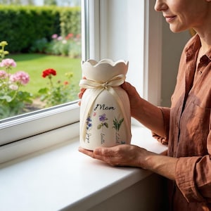 May include: Cream-colored ceramic vase with a scalloped top and a cream-colored ribbon tied around the middle. The vase is decorated with floral illustrations and the word "Mom". The names "Joyce", "Isabella", and "Lilah" are also printed on the vase.