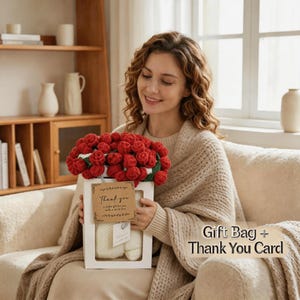 Può includere: Una donna tiene in mano una scatola regalo bianca contenente un bouquet di rose rosse all'uncinetto. La scatola contiene anche un paio di pantofole all'uncinetto bianche e un piccolo biglietto con la scritta "Grazie". Sono visibili anche le parole "Borsa regalo + Biglietto di ringraziamento".