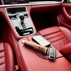 May include: A wooden and silver-toned car accessory with the name "Michell" engraved on it, a smartphone, and a car key fob are displayed on a red leather car interior. The car's gear shift and dashboard are visible in the background.