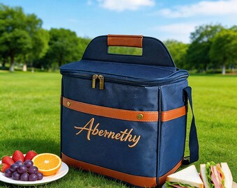 May include: A navy blue insulated cooler bag with brown faux leather trim and a top handle. The name Abernethy is embroidered in gold thread on the front. The bag features a zippered closure and a shoulder strap, designed for carrying food items.