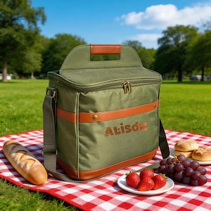 May include: An olive green insulated cooler bag with brown leather accents and the name "Alison" printed on the front. The bag is on a red and white checkered picnic blanket, with food items like strawberries, grapes, and bread.