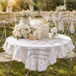 May include: A white round tablecloth with a ruffled edge, featuring the printed text YUKU & JONAS, Jameson, and April.05.2026. The table is decorated with a bouquet of white roses, a white laser-cut lantern, two small gift boxes, and a glass vase.