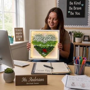 May include: A square white shadow box frame containing a heart-shaped arrangement of paper roses in shades of white and green. The frame features internal LED lighting and white script text that reads Ms. Connie, You are the reason I love learning, Raya.