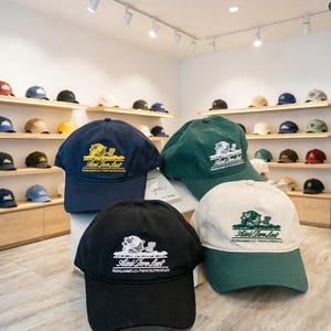 Op de afbeelding: Een display van baseball caps in verschillende kleuren, waaronder marineblauw, zwart, groen en crème. Elke cap heeft een logo met tekst. De caps worden tentoongesteld op planken en een display.
