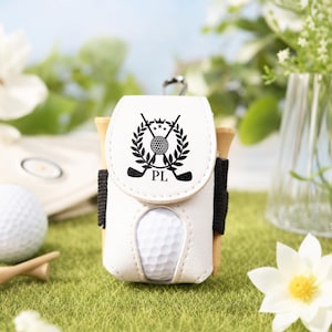 May include: White golf ball holder with a black graphic of crossed golf clubs, a golf ball, and the initials "PL". The holder is attached to a wooden tee holder and holds a white golf ball. The holder is on a green surface.