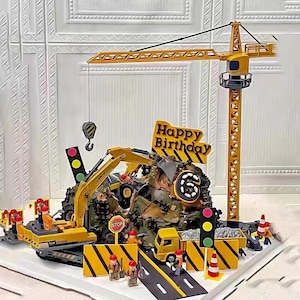 May include: A yellow construction-themed birthday cake with a crane, excavator, and other vehicles. The cake has a "Happy Birthday" sign, traffic lights, and miniature construction worker figures.