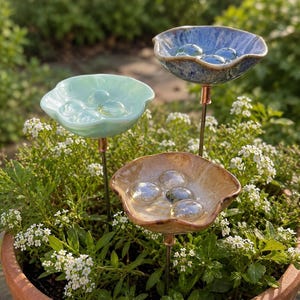 Könnte beinhalten: Drei blumenförmige Gartenstecker aus Keramik in Hellblau, Minzgrün und Hellbraun. Jedes schalenförmige Oberteil hat gewellte Ränder und enthält mehrere klare Glasmurmeln. Die Keramikteile sind auf dünnen Metallstäben mit kupferfarbenen Akzenten montiert.