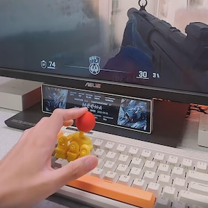 May include: A yellow and red 3D-printed toy robot with a red ball on top, held over a white keyboard with orange keys. The monitor displays a game interface with the number 74 and the word "MECHA".