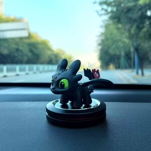 May include: A black dragon figurine with large green eyes and a red emblem on its wing. The figurine is on a black circular base, set against a blurred outdoor background. The dragon is a stylized character.