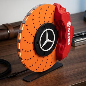 Peut inclure: Un modèle de disque de frein orange décoratif avec un étrier de frein rouge Brembo et un moyeu central noir arborant un logo Mercedes-Benz blanc. Le disque présente de petits points noirs sur sa surface et repose sur une base rectangulaire noire.