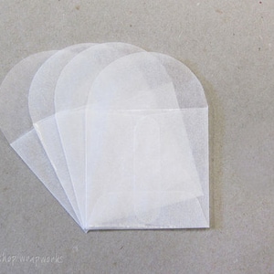 May include: A collection of translucent, white paper envelopes arranged in a fan shape. Each envelope has a rounded top and a rectangular bottom, with a folded flap. The envelopes are stacked on a neutral gray surface.