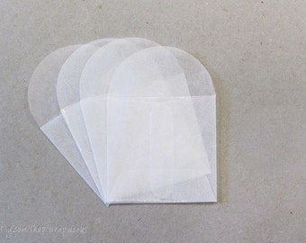 May include: A collection of translucent, white paper envelopes arranged in a fan shape. Each envelope has a rounded top and a rectangular bottom, with a folded flap. The envelopes are stacked on a neutral gray surface.