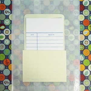 May include: A cream-colored library card pocket with a white card inside, featuring lines for date and issued to. The pocket and card are inside a translucent paper bag, set against a colorful patterned background.