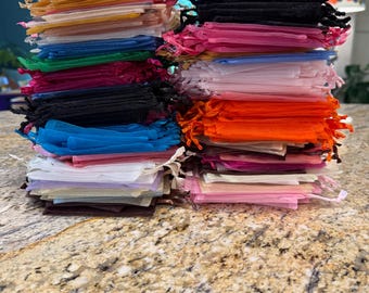 May include: Two large, stacked piles of small, rectangular organza drawstring bags in a variety of colors including black, white, orange, pink, blue, green, yellow, and brown. Each bag features a thin ribbon drawstring closure at the top edge.