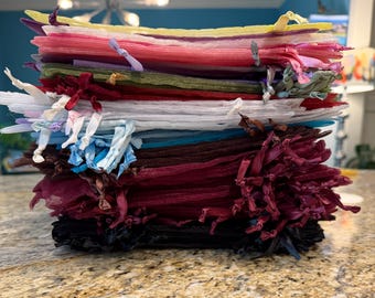 May include: A large stack of rectangular organza drawstring bags in a variety of colors including yellow, pink, red, olive green, white, light blue, dark brown, burgundy, and black, each featuring a matching ribbon tie at the top opening.