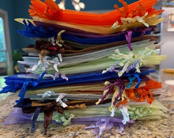 May include: A vertical stack of numerous rectangular organza drawstring gift bags in a variety of colors including orange, blue, brown, light green, and purple. Each sheer fabric pouch features a matching ribbon tie knotted at the top opening.