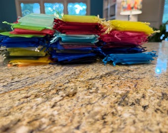 May include: A large collection of small, rectangular organza drawstring bags stacked in multiple piles. The bags are semi-transparent and come in a variety of vibrant colors including red, yellow, blue, green, pink, orange, and purple.