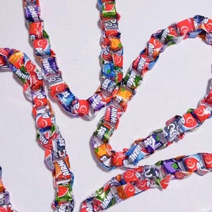 May include: Several strands of colorful candy wrappers are arranged in a wavy pattern against a white background. The wrappers feature various fruit flavors and the Airheads logo. The overall effect is playful and vibrant.