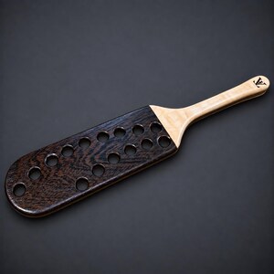 May include: A wooden paddle featuring a dark brown, textured head with fifteen circular cutouts and a light-coloured, smooth handle. The handle displays a small, black, stylized logo at the end, consisting of the letters V and A intertwined.