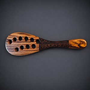May include: A wooden paddle featuring a wide, rounded head with eight circular cutouts arranged in two rows. The wood displays a prominent dark brown and tan grain pattern, transitioning to a darker, solid-toned handle with a smooth, rounded end.