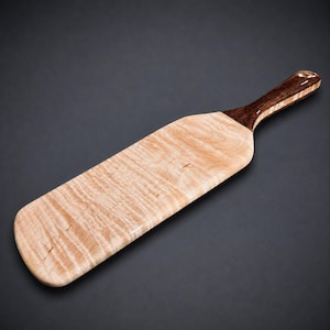 May include: A wooden paddle featuring a light-coloured, wavy-grained rectangular blade and a contrasting dark brown, smooth-finished handle. The wood grain displays a natural, rippled pattern across the surface of the flat, elongated blade.