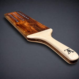 May include: A wooden paddle featuring a dark brown grain on the flat surface with three rows of small metal studs. The handle is made of a lighter-coloured wood and displays a black logo consisting of the letters T and A intertwined at the end.