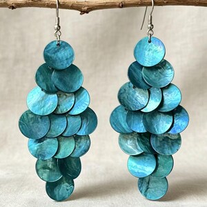 Könnte beinhalten: Ein Paar Ohrhänger mit mehreren überlappenden, kreisförmigen Scheiben aus irisierendem, blau-grünem Muschelmaterial. Die Scheiben sind in einem gestuften, rautenförmigen Muster angeordnet und an silberfarbenen Metallhaken befestigt.