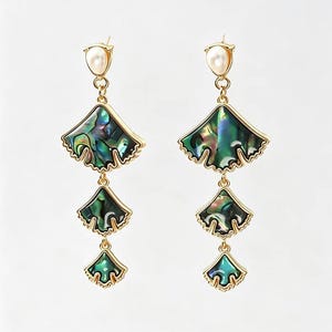 Op de afbeelding: Een paar goudkleurige oorbellen met een witte parel aan de bovenkant, gevolgd door drie aflopende waaiervormige bedels ingelegd met iriserende groene en blauwe abalone schelp, verbonden door kleine goudkleurige metalen ringetjes.