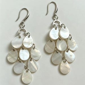 Peut inclure: Une paire de boucles d'oreilles chandelier composées de multiples breloques en nacre blanche en forme de goutte, suspendues à un arceau et un crochet en métal argenté. Chaque boucle présente des pièces en coquillage irisé en cascade.