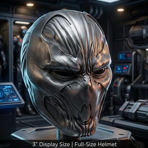 May include: A metallic silver full-size helmet featuring a sculpted, textured surface with deep grooves and sharp, angular contours around the eye sockets and jawline. The helmet is displayed on a stand, with text reading 3" Display Size.