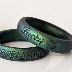 Könnte beinhalten: Zwei grüne und schwarze Silikonringe mit einem keltischen Knotendesign. Ein Ring hat das Wort "Lucky" und ein vierblättriges Kleeblatt eingraviert.