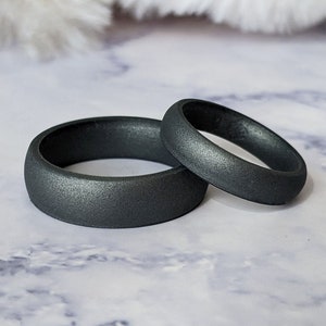 Op de afbeelding: Twee donkergrijze siliconen ringen, een iets groter dan de andere, op een wit marmeren oppervlak.