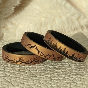 May include: Three silicone rings with different nature-inspired designs. The rings are a dark brown color with a gold-colored finish. The designs are a mountain range, a forest, and waves.