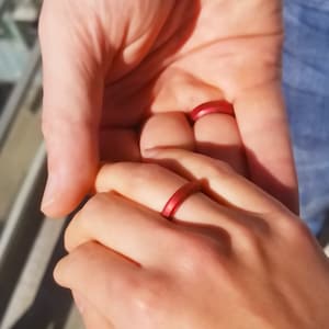 May include: Two hands holding each other, with a red silicone ring on the finger of one hand.