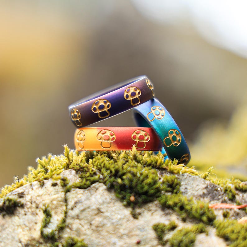 May include: Three colorful rings with mushroom designs. The rings are stacked on top of each other, with the top ring being purple, the middle ring orange, and the bottom ring blue. The rings are set against a blurred background of green moss and a neutral background.