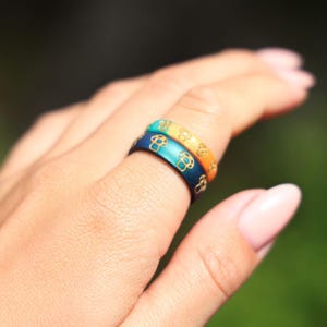 May include: Two colorful rings stacked on a finger. The rings are dark blue, teal, and orange, each with a repeating gold mushroom pattern. The rings are likely made of enamel or resin.